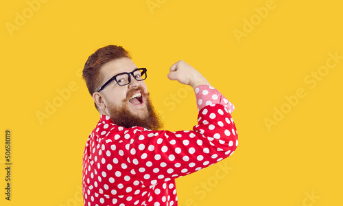 Funny stylish bearded chubby man with smile on face shows showing biceps on yellow background. Joyful crazy redhead millennial man in glasses and red shirt in white peas shows his power. Web banner.