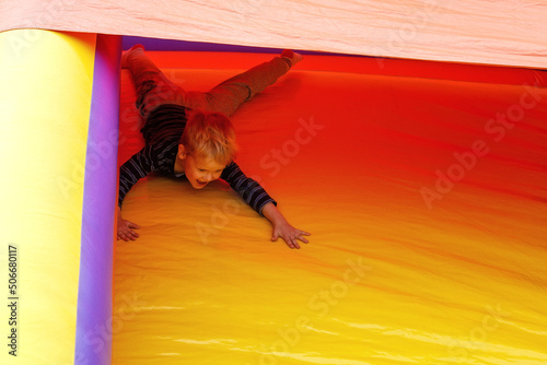 A brave little boy slides down on a red-yellow trampoline at an amusement park.