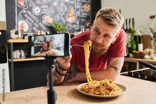 Food blogger eating homemade pasta while filming recipe and mukbang with smartphone on tripod in kitchen