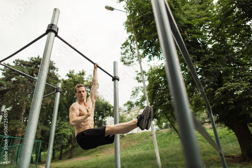 Strong shirtless caucasian athlete performing corner on horizontal bar during street workout on sunny urban sport playground. Healthy lifestyle and street sport concept