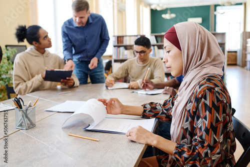 Muslim female immigrant sitting at table looking through sheets of paper with grammar tasks during English lesson