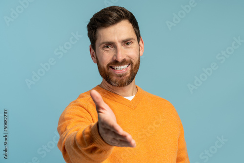 Portrait of bearded man standing with outstretched hand, offering handshake to partner, greeting on job interview. Indoor studio shot isolated on blue background