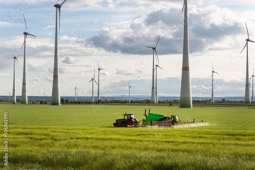 Scenic landscape view big modern tractor machine with sprayer equipment spraying fertilizer in rapeseed agricultural field against windfarm power windmill. Arable land cultivation sustainable energy