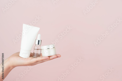 Skincare routine​ for healthy skin. Woman hand holding facial foam tube, serum bottle, moisturizing​ cream on pink background. Beauty​ and cosmetic concept.