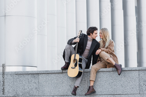 full length of woman in trendy autumn outfit sitting on parapet near man with guitar.