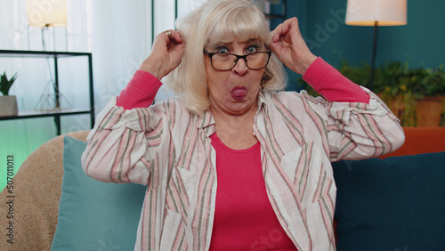 Portrait of happy funny Caucasian senior old grandmother making playful silly facial expressions and grimacing, fooling around, showing tongue. Elderly mature granny woman sitting at home on sofa