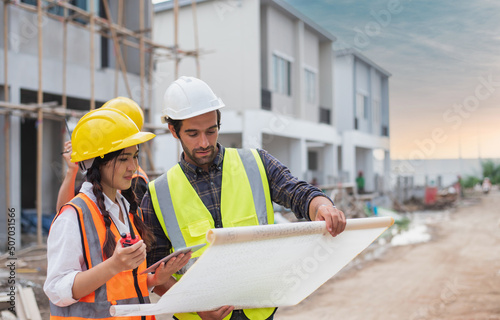 Caucasian males and Asian female builders, architector and engineers with draft plan of building and laptop computer talking on constructing site