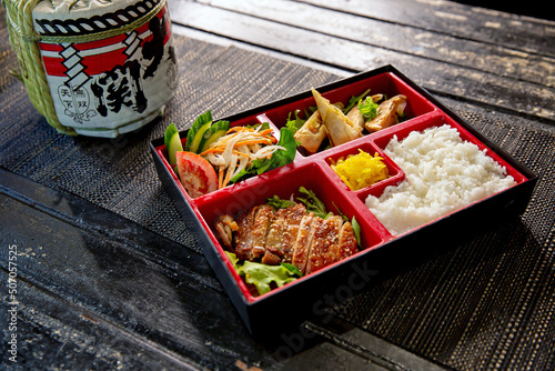 Teriyaki chicken bento with dumplings and salad in the traditional Japanese ramen restaurant, with bento box on and Japanese fonts sake barrel translation: Invincible Great Gate
