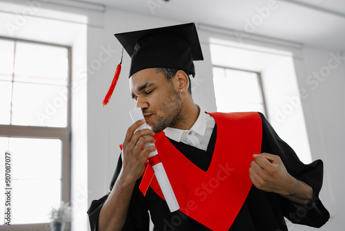 A Black student in graduate gown and square cap who is happy to finish his studies shows his the long-awaited diploma
