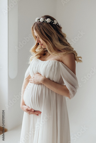 Blonde pregnant woman in a white dress with a wreath on her head in the studio on a white background holds her hand on her stomach