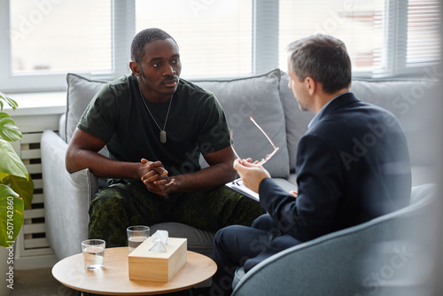 Young African American man wearing military uniform sitting in couch listening to psychotherapist during therapy session