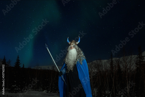 old man Odin with a staff stands in the snow at night against the backdrop of the northern lights