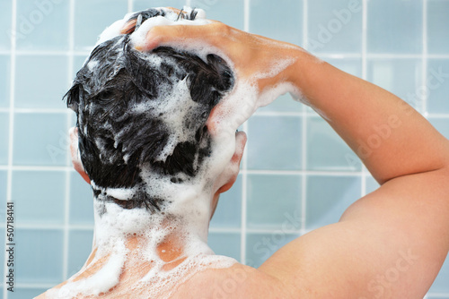 Male hands wash their hair with shampoo and foam on blue background, rear view.
