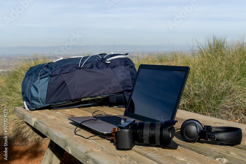 Backpack, computer, camera and headphones on a table in the wilderness - digital nomad concept
