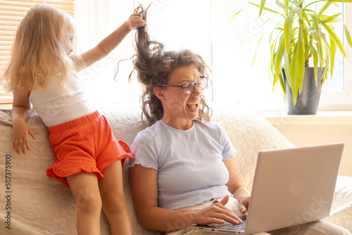 Distant home office during lockdown.Little daughter bothering interfering mother freelancer working using laptop, holding video call with clients, sitting on sofa indoor.Playful girl pulling mom hair