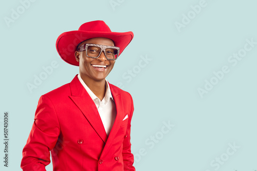 Portrait of happy extravagant dark skinned showman on pastel light blue background. African American man in red jacket, cowboy hat and shiny glasses smiles with snow-white smile near copy space.