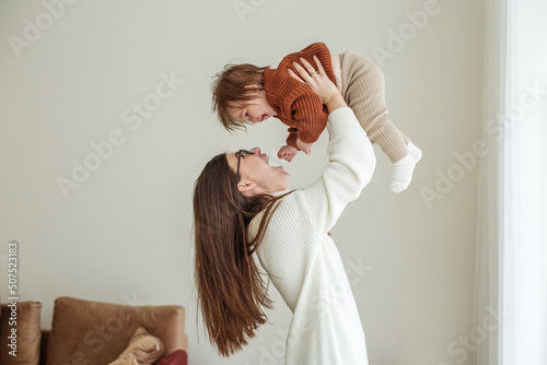 Happy cheerful young mother throws an adorable baby in air, plays, hugs baby with love, care