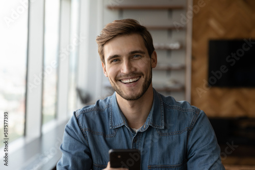 Head shot of happy young millennial man holding smartphone, looking at camera, smiling, laughing. Portrait of positive customer, smartphone user satisfied with online app, virtual service work