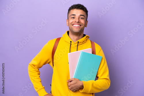 Young student Brazilian man isolated on purple background posing with arms at hip and smiling