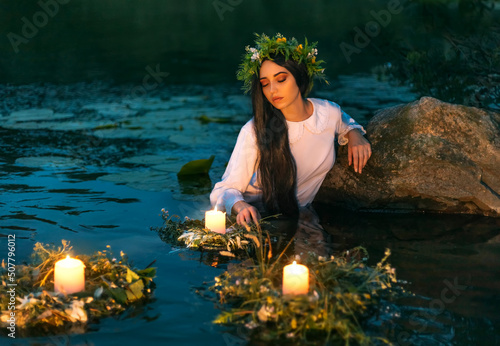 Slavic woman nymph stands in water herbal wreath floa candles burning. Fantasy girl mermaid. White long wet dress. Summer night green grass tree river. pagan holiday Ivan Kupala divination maidens