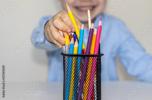 joyful kid takes a pencil from metal net box pen holder in front of him. back to school concept.cute little boy in blue shirt.elementary school or homeschooling, distance learning. colorful pencils