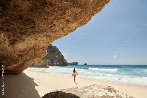 Young beautiful girl in a yellow swimsuit is sunbathing while standing on a tropical beach with white sand and turquoise water. Vacation on Diamond Beach in Nusa Penida Bali Indonesia
