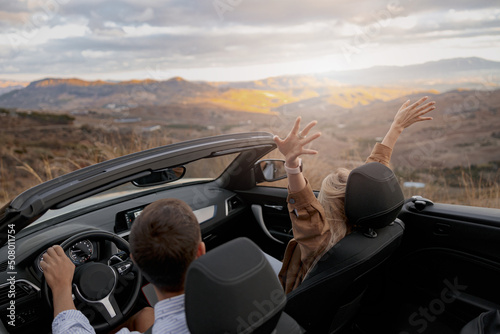 Rear of couple man and woman traveling in convertible car by mountainside enjoying panorama view