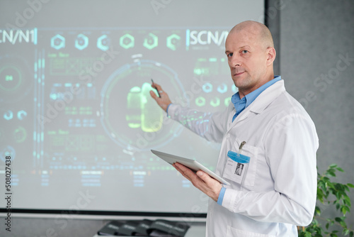 Portrait of mature male doctor in white coat with digital tablet pointing at projector with medical charts at conference