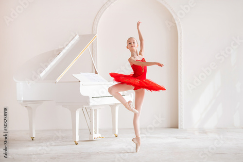 A cute little girl dreams of becoming a professional ballerina. In a white room, next to a piano, a girl in a bright red tutu is dancing on pointe shoes. Vocational school student.