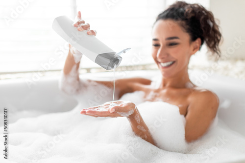 Beautiful young woman taking bubble bath, pouring shampoo into her hand, enjoying home spa procedure, selective focus