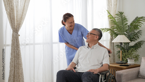 Friendly taiwanese woman nursing aide putting hands on disabled grandfather’s shoulders while talking to him at home. he sits on a wheelchair and has smile on face