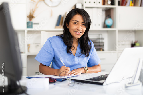 Smiling peruvian female therapist working with case histories on laptop in modern medical office