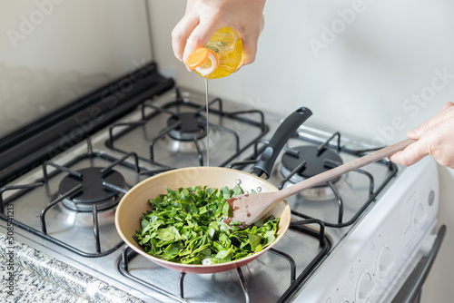 Female hands cooking ora-pro-nobis in frying pan. Pouring oil and wooden spoon. Pereskia aculeata is a popular vegetable in parts of Brazil