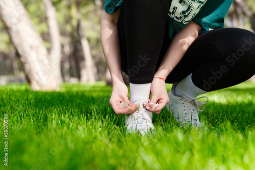 Young caucasian woman wearing green tee standing on city park, outdoor tying lace running shoes getting ready for run. Jogging girl exercise motivation health and fitness.