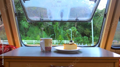 View from caravan window on road and forest with cup of hot coffee or tea and cake on table. Scenic spruce woodland view from RV camper van dinette window. Vacation, travel motorhome getaway concept.