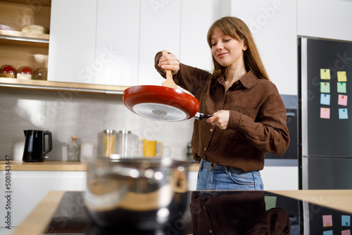Young woman at the stove stirring food in a frying pan with a spatula. Housewife cooks in the kitchen