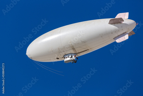 A white blimp without any markings, a blank canvas or banner space with a blue sky in the background. A lighter than air ship flying high with room to put your own ad.