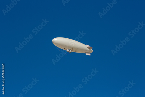 A white blimp without any markings, a blank canvas or banner space with a blue sky in the background. A lighter than air ship flying high with room to put your own ad.