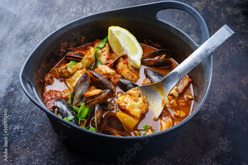Modern style traditional Spanish seafood zarzuela de pescado with fish, king prawns and blue mussels served in red sauce as close-up in design pot