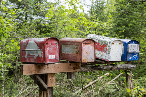 Closeup selective focus view of four old rusty mailboxes on a totting wooden post with blurry trees in background in rural village. Copy space above.