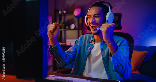 Happy asia guy gamer wear headphone participation play video game colorful neon lights computer in living room at night modern house. Esport streaming game online, Home quarantine activity concept.