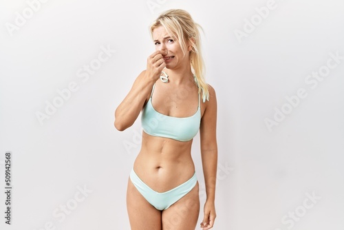 Young caucasian woman wearing bikini over isolated background smelling something stinky and disgusting, intolerable smell, holding breath with fingers on nose. bad smell