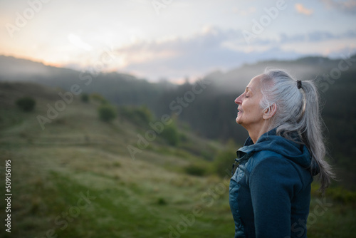 Senior woman doing breathing exercise in nature on early morning with fog and mountains in background.