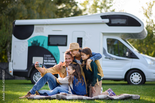 Happy young family with two children ltaking selfie with caravan at background outdoors.
