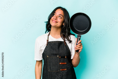 Young hispanic cooker woman holding frying pan isolated on blue background dreaming of achieving goals and purposes