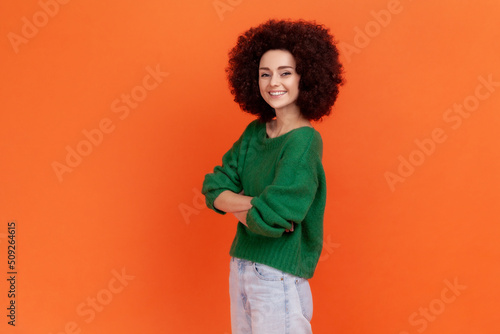 Side view portrait of smiling woman with Afro hairstyle wearing green casual style sweater standing with folded hands and looking at camera. Indoor studio shot isolated on orange background.