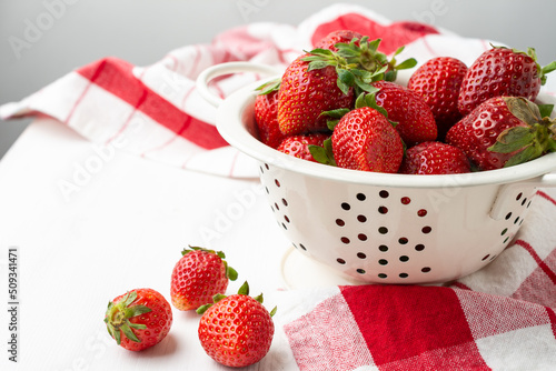Aerial view of strawberries in a white colander on a white table with a red kitchen towel, white background, horizontal