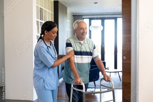 Biracial female physiotherapist assisting happy caucasian senior man in walking with walker at home