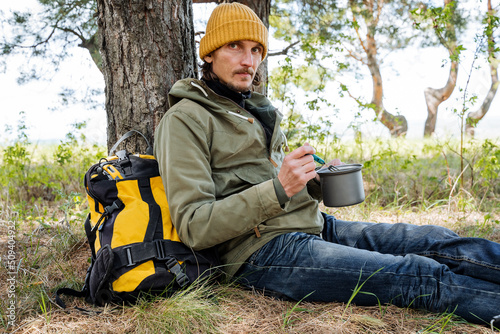 A man sits on the ground in the forest, a tourist on a hike leans his back against a tree, a man has breakfast in nature, survival in the wild, a vacation with a backpack, warm clothes.