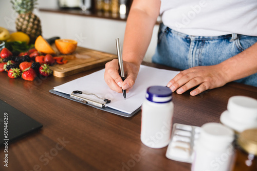 Closeup cropped photo of plump plus size woman counting calories, writing on clipboard healthy food, medicines, pills containers. Nutrition concept